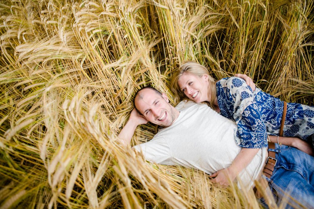 a photograph of a couple lying in a cornfield. The man is looking at the camera and smiling, while the woman is smiling at him.