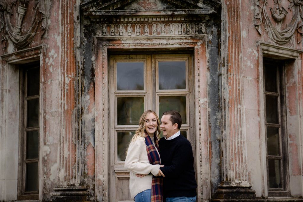a couple stand in the doors of the roman temple at larmer tree gardens. they are laughing.