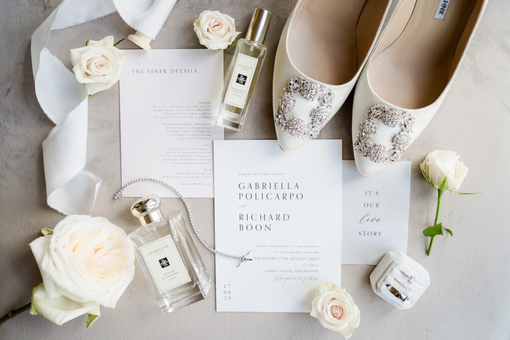 flat lay photograph of wedding details at Caswell House featuring Jo Malone purfume, Manolo Blahnik shoes, and wedding rings surrounded by white roses