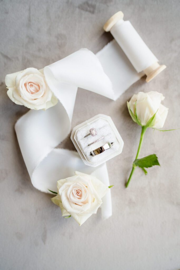 flat lay photograph of wedding rings in a white velvet box with a spool of white silk ribbon and white roses