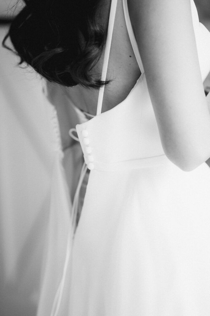 the back of a bride's Suzanne Neville wedding dress is open, you can see the buttons and spaghetti straps
