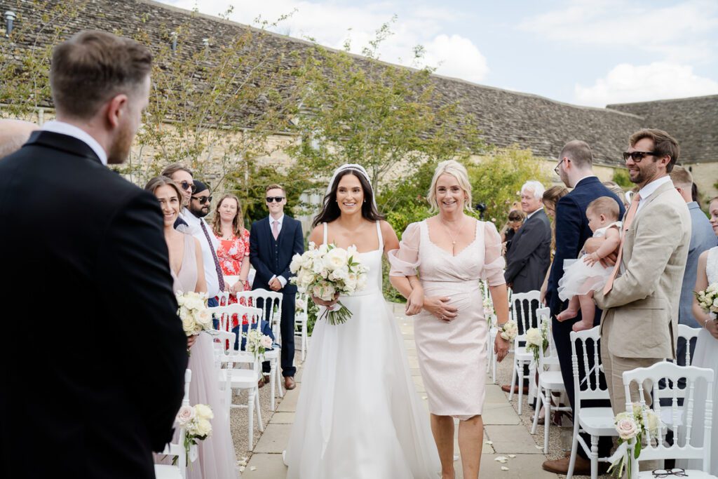 a Bride is walked down the aisle by her mother in the gardens of Caswell House. The guests look on and her Groom is in the foreground.